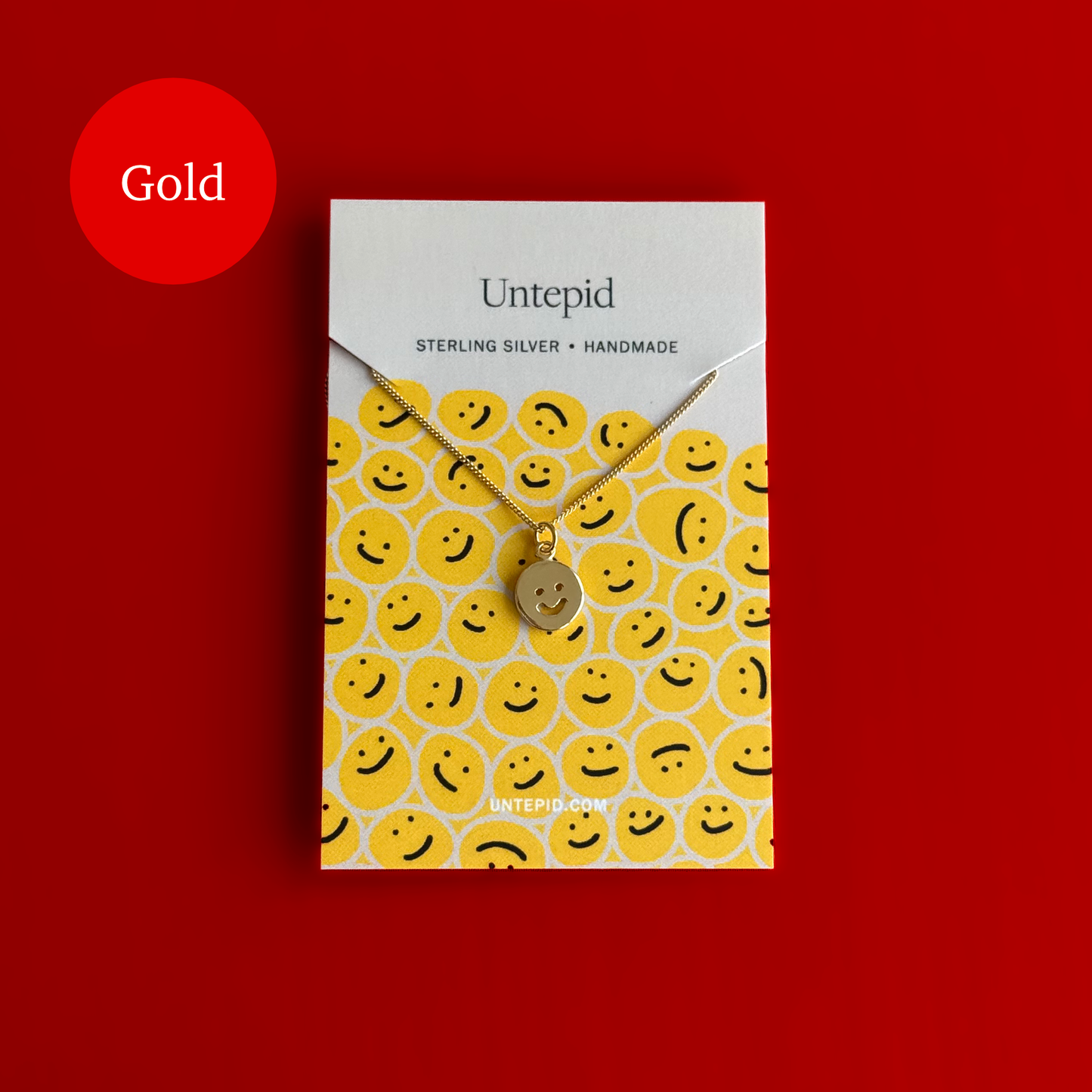 A gold-toned Untepid necklace with a smiley face charm, displayed on its branded yellow and white backing card against a solid red background. A red circular badge in the top-left corner reads 'Gold'.